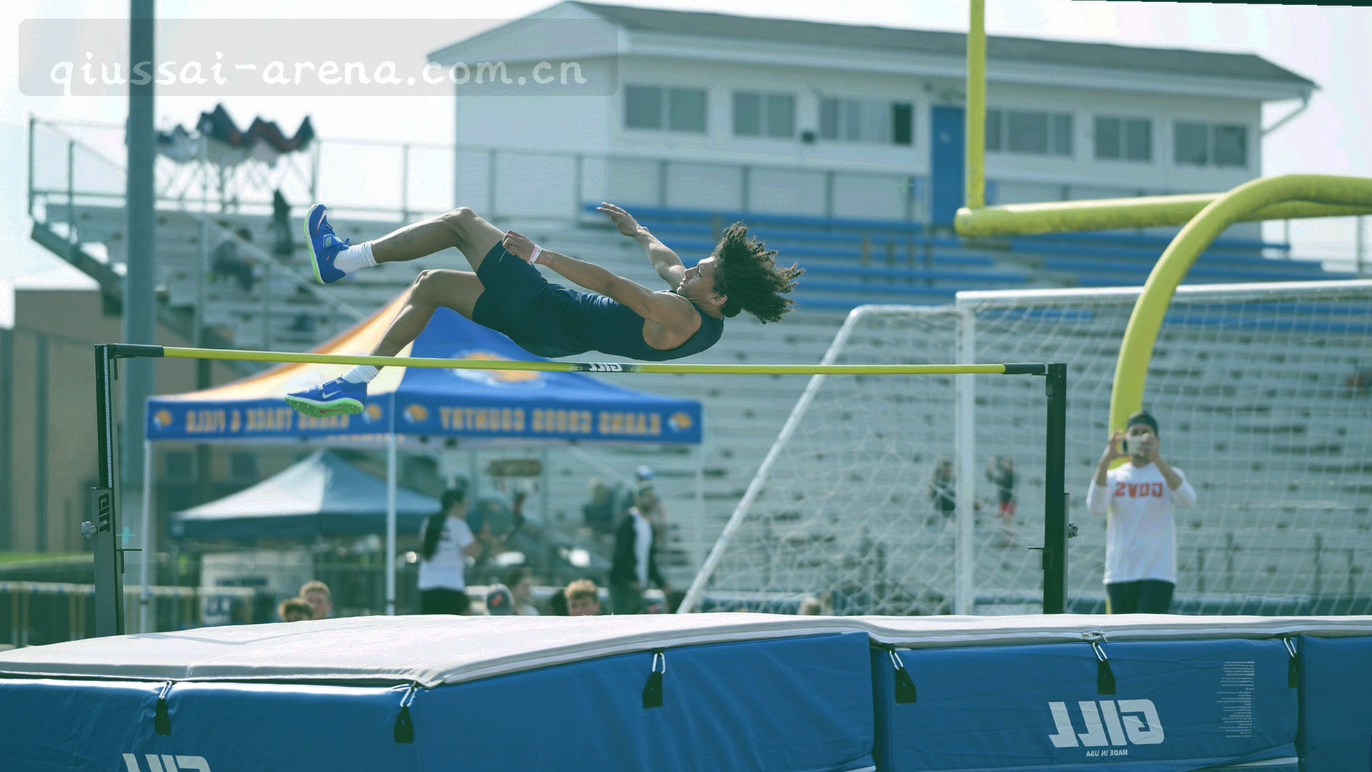 夜间球场俯拍，灯光照亮绿色草皮与看台阴影，电子计分牌的光晕悬在场地一角